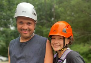 A young counselor and an adult camper with disabilities are wearing hardhats and climbing gear