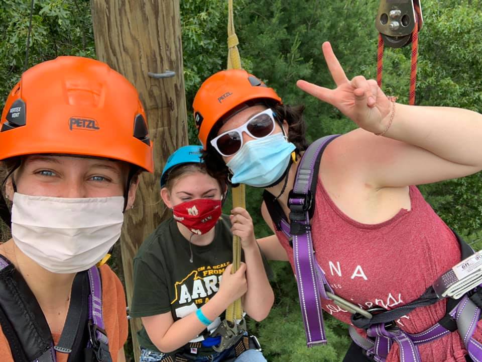 Two adults in climbing gear wearing masks help a kid camper in climbing gear