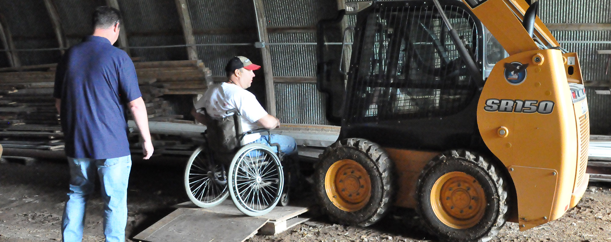 Two farmers, one in a wheelchair, looking at a piece of machinery inside an out building